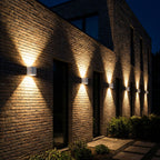 Modern rectangular outdoor wall lights mounted on a brick building at night, casting warm light beams downward in a row along the facade.